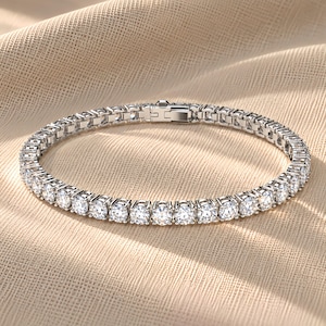 May include: A silver-toned tennis bracelet featuring a continuous row of round, clear, faceted stones set in a metallic frame. The bracelet includes a secure box clasp mechanism with a hinged closure, displaying a uniform design throughout the piece.