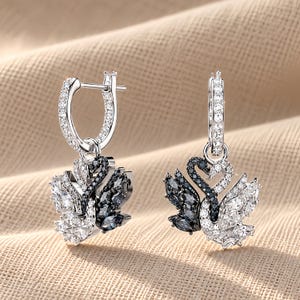 May include: A pair of silver-toned hoop earrings featuring dangling swan-shaped pendants. Each swan is encrusted with clear and black crystals in a marquise and round cut. The hoops are also adorned with small clear crystals along the front surface.