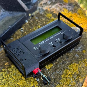 May include: A black QRP Labs QMX Transceiver featuring a rectangular digital display, two circular control knobs, and a series of small buttons. The device has a textured black casing with a small red and green electronic component attached to the side.