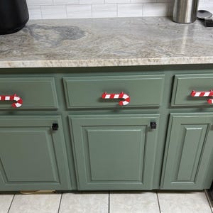 May include: Three red and white striped candy cane shaped drawer pulls attached to sage green kitchen cabinets. The cabinet hardware features a classic curved hook design with alternating red and white diagonal stripes, providing a festive accent.