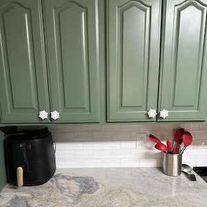 May include: Sage green kitchen cabinets featuring white snowflake-shaped knobs. A black air fryer sits on the counter next to a stainless steel utensil holder filled with red silicone kitchen tools against a white subway tile backsplash.