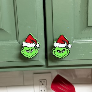 May include: Two cabinet knobs shaped like the head of a green character with a furrowed brow and a smirk, wearing a red and white Santa hat. The knobs are mounted on sage green cabinet doors, featuring a distinct black outline and textured finish.
