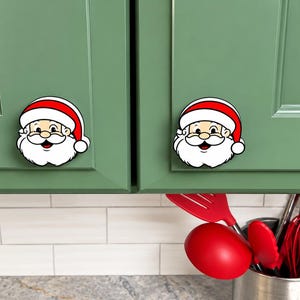 May include: Two decorative cabinet knobs shaped like Santa Claus faces. Each knob features a smiling Santa with a white beard, a red and white hat, and a black outline. The knobs are mounted on green cabinet doors, providing a festive holiday accent.