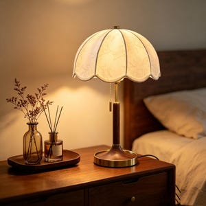 May include: A table lamp with a scalloped white fabric shade featuring a subtle floral pattern, a dark wood grain stem, and a brass base. A pull chain switch hangs from the shade. Beside it sits a glass vase with dried flowers and a reed diffuser.