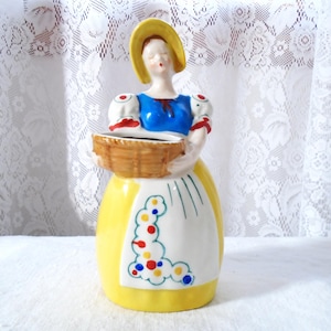 May include: A ceramic figurine of a woman in a yellow dress and a yellow hat, holding a brown basket. The figurine has a blue top and a white apron with colorful polka dots. The background is a white lace curtain.