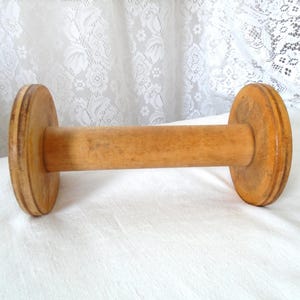 May include: A vintage wooden spool featuring a cylindrical central shaft and two circular end discs. The wood displays a natural light brown finish with visible grain patterns and signs of wear, including minor surface imperfections and texture.