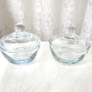 May include: Two clear glass candy dishes with lids. Each dish has a rounded bowl shape and a clear, round knob on top of the lid. The dishes are sitting on a white surface, with a white lace curtain in the background.