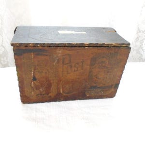 May include: A rectangular wooden box with a dark brown finish and a hinged lid. The front panel features the word Post in a stylized font, flanked by two faded graphic illustrations of figures. The edges of the box show a textured, notched design.
