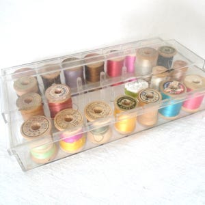 May include: A clear plastic sewing box filled with various spools of thread. The spools are made of wood and contain threads in a variety of colors, including pink, blue, yellow, and brown. The box is rectangular and has a clear lid.