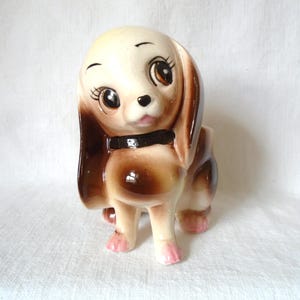 May include: A ceramic dog figurine with a cream-colored head and brown ears, body, and accents. The dog has large, expressive eyes, a black collar, and pink paws. The figurine is a decorative item.