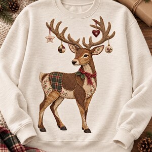 May include: A cream-colored crewneck sweatshirt featuring a large, central graphic of a reindeer. The reindeer is depicted with a patchwork-style body in shades of brown, green, and red, wearing a red scarf and adorned with hanging ornaments.