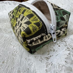 May include: A small, open, rectangular zippered pouch. The pouch features a repeating geometric pattern in shades of green, black, and cream. The zipper is white, and the interior lining is a cream-colored fabric with small white polka dots.