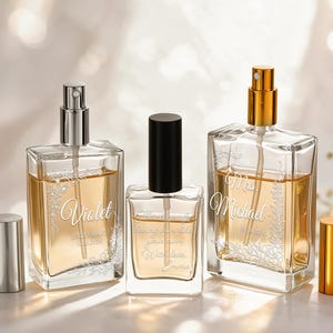 May include: Three rectangular glass perfume bottles filled with amber liquid. The left bottle has a silver pump and cap with the name Violet engraved. The center bottle has a black cap. The right bottle has a gold pump and cap with the name Michael engraved.