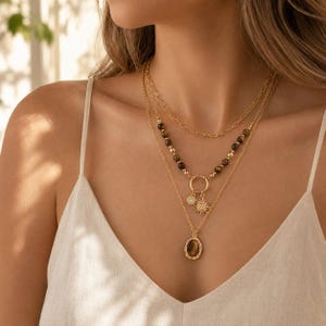 May include: A layered gold-tone necklace set featuring a top paperclip chain, a middle strand with brown tiger eye beads and a circular pendant with sun and crystal charms, and a bottom strand with an oval tiger eye stone in a textured frame.
