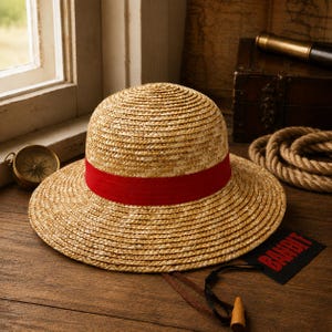 May include: A straw hat with a rounded crown and a wide brim, featuring a vibrant red fabric band around the base. A black rectangular tag with the red text "BANDIT" is attached to the hat by a thin cord with a small wooden toggle at the end.