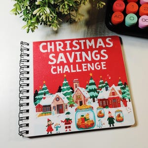 May include: A square spiral-bound notebook with a red cover featuring the white text CHRISTMAS SAVINGS CHALLENGE. The cover illustration depicts a snowy village with houses, pine trees, and characters including Santa Claus and jars filled with gold coins.