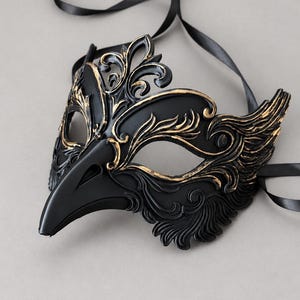 Può includere: Una maschera da ballo in maschera nera con un becco appuntito e dettagliati rilievi a volute evidenziati in oro. La maschera presenta due aperture per gli occhi ed è fissata a lunghi nastri di raso nero per legarla al capo.
