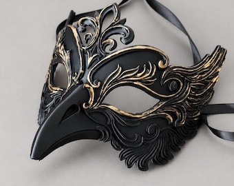 May include: A black masquerade mask with a pointed beak and intricate raised scrollwork detailing highlighted in gold. The mask features two eye openings and is attached to long black satin ribbons for securing it around the head.