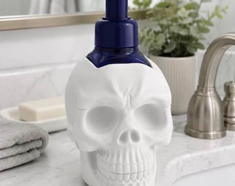 May include: A white skull-shaped soap dispenser featuring a dark blue plastic pump mechanism. The skull is molded with detailed eye sockets, a nasal cavity, and teeth, serving as the base for the liquid soap container.