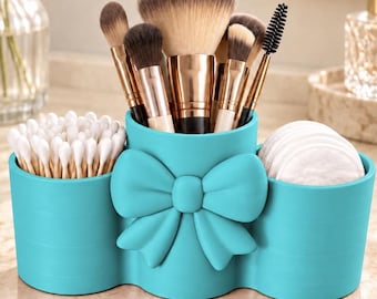 May include: A turquoise makeup organizer with three cylindrical compartments and a prominent bow detail on the center section. The organizer holds various makeup brushes with metallic handles, a cluster of white cotton swabs, and white cotton pads.