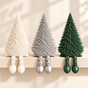 Può includere: Tre figure decorative a forma di cono con fogliame testurizzato simile ad aghi nei colori crema, argento e verde scuro. Ogni figura presenta due gambe articolate e pendenti con piedi arrotondati per sedersi su un bordo.