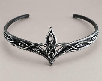 May include: A silver-toned metal circlet featuring a weathered, dark-accented finish. The headpiece has a central pointed element with an intricate, interwoven knotwork design, flanked by symmetrical, vine-like branches extending along the band.