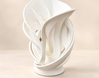 May include: A white ceramic sculpture featuring a series of fluid, overlapping, and curved ribbon-like forms that rise from a circular base. The object has a matte finish and an intricate, open structure with multiple layered segments.