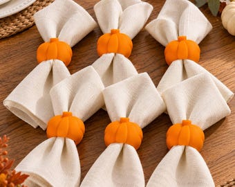 May include: A set of six off-white fabric napkins, each secured with a bright orange pumpkin-shaped napkin ring. The napkins are folded into a pointed shape, and the pumpkin rings feature a smooth, matte finish with visible vertical ridges.