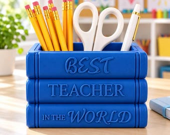 May include: A blue desk organizer shaped like a stack of three books with the embossed text BEST TEACHER IN THE WORLD. The organizer holds several yellow pencils with pink erasers, a pair of white scissors, and a white pen with a gold clip.