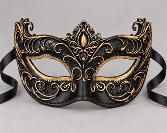 May include: A black masquerade mask featuring intricate gold-colored filigree detailing across the surface. The mask has a central raised crest, almond-shaped eye openings, and two black satin ribbons attached to the sides for securing.
