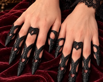 May include: A set of black claw-style finger rings and matching band rings featuring intricate filigree patterns and heart-shaped cutouts. The pointed, elongated nail covers extend over the fingertips, while the bands sit at the base of the fingers.