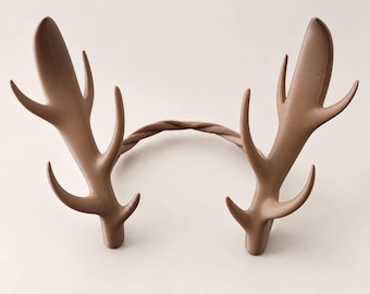 May include: A brown headband featuring two symmetrical deer antler attachments. The antlers have a smooth, matte finish with multiple points extending upwards and outwards. The headband itself has a twisted, rope-like texture connecting the two antlers.