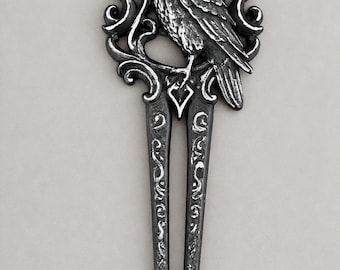 May include: A silver-toned metal hair fork featuring a detailed raven perched within an oval frame. The frame is decorated with swirling scrollwork and a crescent moon at the top. Two long, slender prongs extend from the base of the raven design.