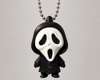 May include: A small, stylized figurine pendant featuring a white ghost-like mask with hollow eyes and an open mouth, attached to a black hooded robe. The figure is suspended from a silver-toned metal ball chain necklace, creating a distinct accessory.