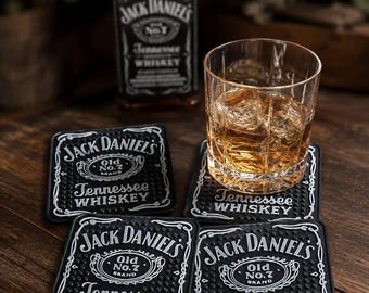 May include: Three square black rubber bar coasters featuring the white Jack Daniel's Tennessee Whiskey logo and a textured nub surface, accompanied by a glass of amber liquid with ice cubes and a bottle of Jack Daniel's Tennessee Whiskey.