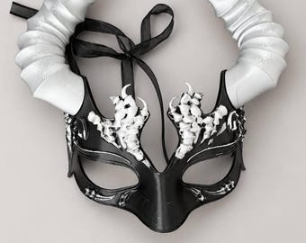 May include: A black masquerade mask featuring two large, curved silver horns extending upward. The mask is decorated with silver, bone-like filigree accents above the eye openings and includes black ribbon ties for securing the item in place.