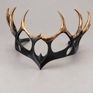 Può includere: Un copricapo con una base nera opaca e una struttura appuntita a forma di corona. Le sezioni superiori delle sporgenze curve, simili a corna, sono rifinite con vernice oro metallizzato, creando un netto contrasto con la struttura scura.