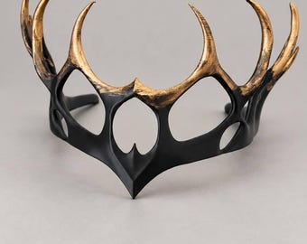May include: A headpiece featuring a matte black base with a pointed, crown-like structure. The upper sections of the curved, antler-like protrusions are finished with a metallic gold paint, creating a distinct contrast against the dark frame.