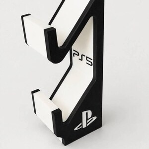 May include: A black and white vertical gaming controller stand featuring a cutout design. The stand includes the text PS5 printed in black and the PlayStation logo symbol at the base. The structure has two distinct cradles for holding hardware.