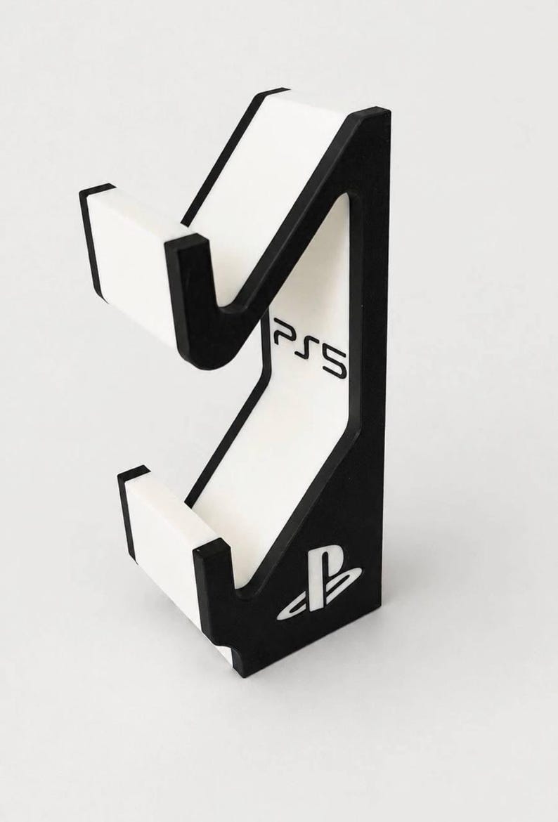 May include: A black and white vertical gaming controller stand featuring a cutout design. The stand includes the text PS5 printed in black and the PlayStation logo symbol at the base. The structure has two distinct cradles for holding hardware.