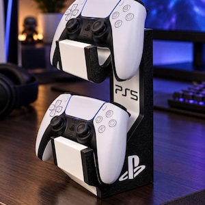 May include: A black and white vertical controller stand holding two white and black game controllers. The stand features the text PS5 and the PlayStation logo printed on the side. The structure has two tiered cradles designed to support the controllers.