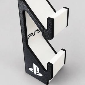 May include: A black and white controller stand featuring a textured black frame with white accents. The stand includes a white vertical panel with the text PS5 printed in black and a white PlayStation logo emblem at the base of the structure.