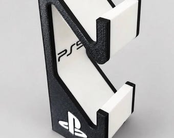 May include: A black and white controller stand featuring a textured black frame with white accents. The stand includes a white vertical panel with the text PS5 printed in black and a white PlayStation logo emblem at the base of the structure.