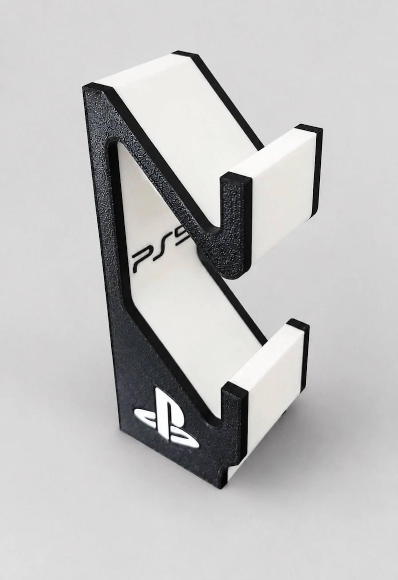 May include: A black and white controller stand featuring a textured black frame with white accents. The stand includes a white vertical panel with the text PS5 printed in black and a white PlayStation logo emblem at the base of the structure.