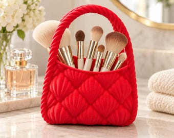 May include: A bright red silicone makeup brush holder shaped like a small handbag with a quilted shell pattern and a braided handle. The container is filled with various makeup brushes featuring gold-toned metal ferrules and soft bristles.