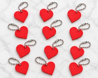 May include: A collection of twelve bright red heart-shaped tags, each featuring a smooth, flat surface and a silver-toned metal ball chain connector attached through a small hole at the top of each heart, arranged in a grid pattern.