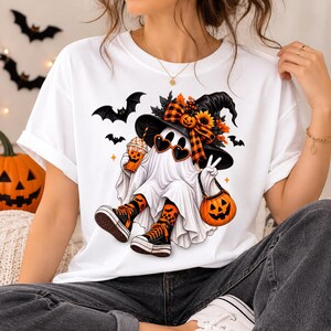 Pode incluir: Uma t-shirt branca com o gráfico de um fantasma usando um chapéu de bruxa preto com fitas xadrez laranja, óculos de sol em forma de coração e ténis. O fantasma segura uma bebida de abóbora e um balde de doces em forma de abóbora.
