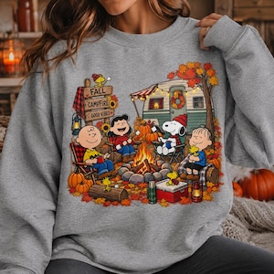 Puede incluir: Una sudadera gris jaspeado con un estampado gráfico de personajes de Peanuts acampando. El diseño muestra a Charlie Brown, Lucy, Snoopy y Linus alrededor de una fogata con una caravana, calabazas y un letrero que dice FALL CAMPFIRE GOOD VIBES.