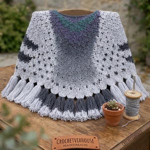 Puede incluir: Un poncho de ganchillo con un degradado de hilo gris claro, gris oscuro y verde azulado, con un dobladillo festoneado y largos flecos. Se ve una etiqueta de cuero con la inscripción CROCHETVIAHOUSE HANDMADE, un carrete de hilo y una suculenta.