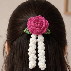 Könnte beinhalten: Ein gehäkeltes Haaraccessoire mit einer leuchtend pinken Rose, zwei kleinen grünen Blättern an der Basis und zwei langen, herabhängenden weißen, strukturierten Strängen. Das florale Design ist aus Garn mit sichtbaren Maschen gefertigt.